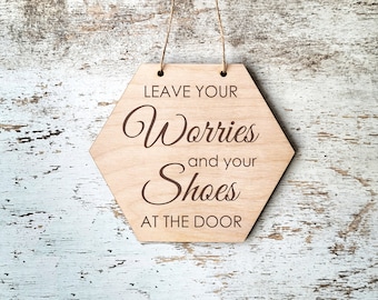 May include: A light-colored wooden sign in a hexagonal shape, with the words "LEAVE YOUR Worries and your Shoes AT THE DOOR" engraved in a dark brown script. The sign hangs from a natural twine string.