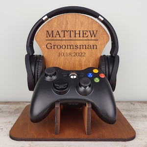 Può includere: Un supporto in legno con una targa personalizzata che recita "MATTHEW Groomman 10.18.2022". Il supporto tiene un auricolare da gioco nero e un controller Xbox nero.