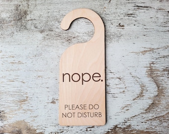 Puede incluir: Un colgador de puerta de madera con el texto "nope." y "Por favor, no molestar" en letras negras.