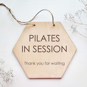 Könnte beinhalten: Ein sechseckiges Holzschild mit dem Text "PILATES IN SESSION" und "Thank you for waiting" in braunen Buchstaben.