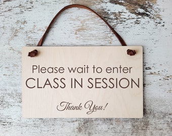 May include: A wooden sign with a brown leather strap hanging from the top. The sign reads "Please wait to enter CLASS IN SESSION Thank You!"