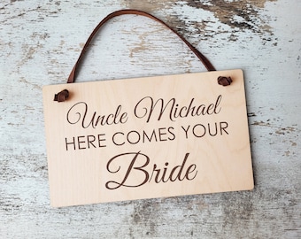 May include: A rectangular light-colored wooden sign featuring the dark brown engraved text "Uncle Michael HERE COMES YOUR Bride". The sign includes two holes at the top with a brown leather cord attached for hanging, creating a simple and functional display.