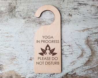 Peut inclure: Panneau de porte en bois clair avec une partie supérieure incurvée. Il porte l'inscription "YOGA EN COURS" au-dessus d'un motif de fleur de lotus et de silhouette, et "MERCI DE NE PAS DÉRANGER" en dessous.