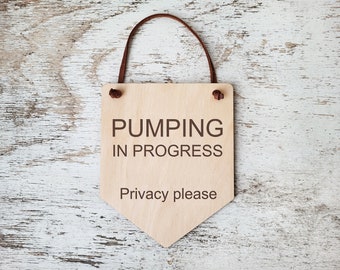 Può includere: Un cartello di legno con un cinturino in pelle marrone è appeso su uno sfondo di legno sbiancato. Il cartello recita "Pumping in Progress, Privacy please".