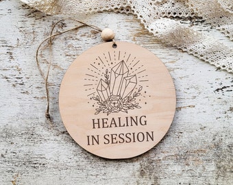 May include: A round wooden sign with a crystal design and the text "HEALING IN SESSION".
