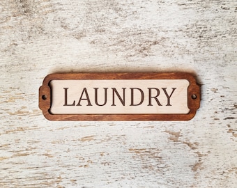 Op de afbeelding: Een houten bord met een bruin frame en een witte achtergrond. Het bord heeft de tekst "LAUNDRY" in bruine letters.