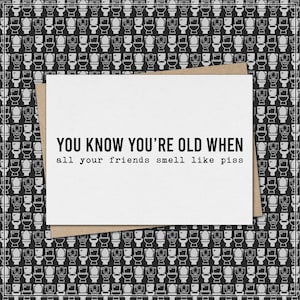 May include: A white greeting card with the text "YOU KNOW YOU'RE OLD WHEN all your friends smell like piss" on a brown envelope, set against a black and white toilet pattern background.
