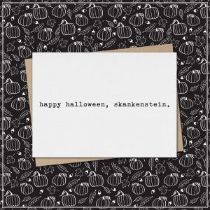 May include: A white rectangular greeting card with the black typewriter-style text "happy halloween, skankenstein." printed in the centre. The card is placed on a black background featuring a repeating white line-art pattern of pumpkins and leaves.