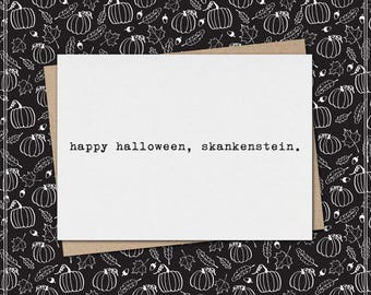 May include: A white rectangular greeting card with the black typewriter-style text "happy halloween, skankenstein." printed in the center. The card is placed on a black background featuring a repeating white line-art pattern of pumpkins and leaves.