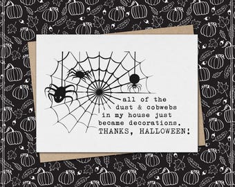 May include: A white greeting card featuring a black ink illustration of a spider web with two spiders. The text reads: all of the dust & cobwebs in my house just became decorations. THANKS, HALLOWEEN! The card is placed on a brown envelope.