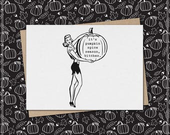 May include: A white greeting card featuring a black ink illustration of a person holding a large pumpkin. The pumpkin has the text 'it's pumpkin spice season, bitches.' printed on its surface. The card is placed on a black background with pumpkins.