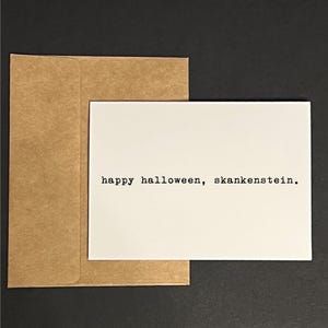 May include: A white rectangular greeting card featuring the black typewriter-style text "happy halloween, skankenstein." printed in the centre, accompanied by a matching brown kraft paper envelope positioned behind the card.