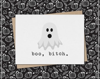May include: A white greeting card featuring a light gray ghost illustration centered above the black text "boo, bitch." The card is placed against a black background patterned with white line drawings of pumpkins, leaves, and acorns.