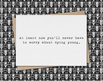 Puede incluir: Una tarjeta de felicitación blanca con el texto "at least now you'll never have to worry about dying young." se encuentra sobre un sobre marrón y un fondo con un patrón de inodoros en blanco y negro. La tarjeta es un regalo humorístico y sarcástico.