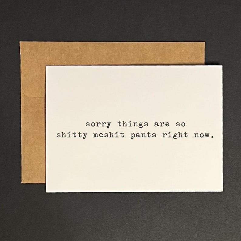 May include: A white greeting card with the text "sorry things are so shitty mcshit pants right now" in black font, placed on a brown envelope. The card and envelope are on a dark surface.