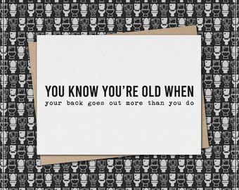 Può includere: Un biglietto di auguri bianco con la scritta "YOU KNOW YOU'RE OLD WHEN your back goes out more than you do." Il biglietto è su una busta marrone. Lo sfondo presenta un motivo di illustrazioni di servizi igienici in bianco e nero.