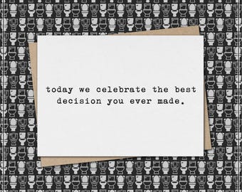Puede incluir: Una tarjeta de felicitación blanca con el texto "today we celebrate the best decision you ever made." está colocada sobre un sobre marrón. El fondo presenta un patrón repetido de ilustraciones de inodoros en blanco y negro.
