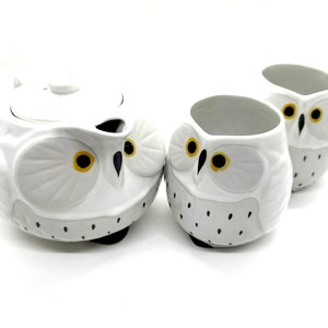 May include: A white ceramic owl-shaped tea set with a teapot and two mugs. The teapot has a lid and spout. The owl's eyes are yellow and black, with gray accents. The mugs have the same owl design. The set is on a white background.