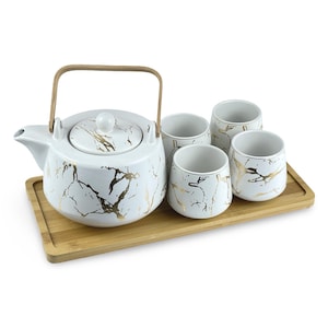 May include: A white ceramic teapot with a gold marble design and a wooden handle, along with four matching teacups, all on a bamboo tray.