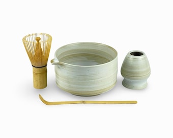May include: A white ceramic matcha bowl with a bamboo whisk, a bamboo spoon, and a white ceramic matcha scoop. The bowl has a light brown glaze and a spout.