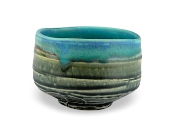 May include: A handmade ceramic bowl with a unique, organic shape. The bowl features a gradient of colors, transitioning from a deep teal at the base to a vibrant turquoise and blue at the rim. The textured surface adds to its artisanal appeal.
