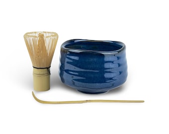 May include: A matcha tea set featuring a deep blue ceramic bowl, a bamboo whisk, and a bamboo scoop. The bowl has a textured surface. The whisk is made of many thin bamboo pieces. The scoop is long and curved.
