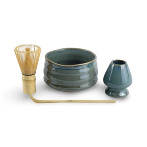 May include: A green ceramic matcha bowl, whisk, and teacup. The bowl has a textured, wavy design. The whisk is made of bamboo and has a wooden handle. The teacup is a traditional Japanese style.