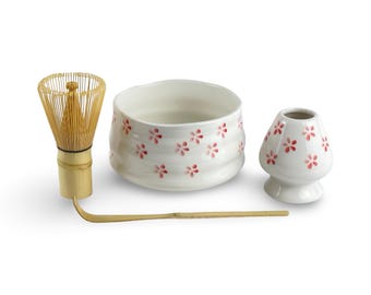 May include: A white ceramic matcha bowl with a pink floral design, a bamboo matcha whisk, and a white ceramic matcha scoop.