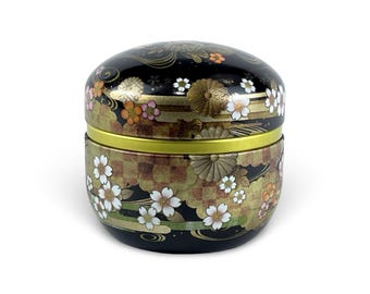 May include: A black, round container with a lid, adorned with floral and geometric patterns in gold, white, and orange. The container has a gold band around the middle and is likely used for storage or display.