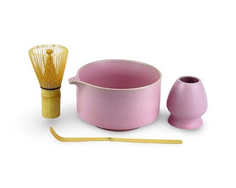 May include: A pink ceramic matcha bowl, whisk, and cup set. The bowl has a speckled finish and a spout. The whisk is made of bamboo. The cup is also speckled pink.