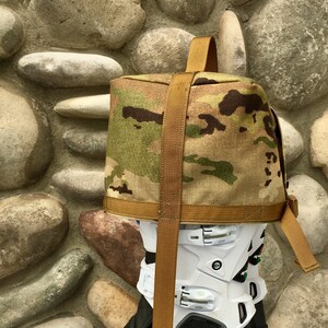 May include: A pair of black motorcycle boots with a white and gray ankle brace, secured with a brown strap and a camouflage fabric bag.