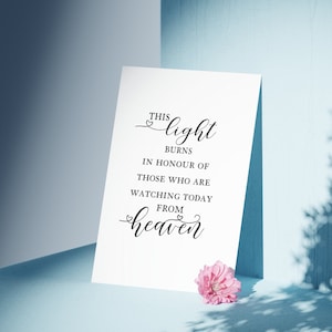 May include: A white card with black text that reads "This light burns in honour of those who are watching today from heaven." The card is sitting on a light blue surface with a pink flower.