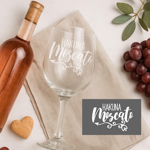 Peut inclure: Un verre à vin transparent avec l'inscription "HAKUNA Moscato" en blanc. Une bouteille de vin rosé, un bouchon en forme de cœur et une grappe de raisin sont également présents. Le verre est posé sur une serviette en lin beige.