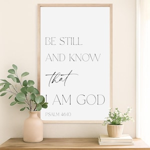 May include: A framed print with the text "BE STILL AND KNOW THAT I AM GOD" in black. The artwork is displayed on a wooden table, accompanied by a vase of greenery and a small potted plant.