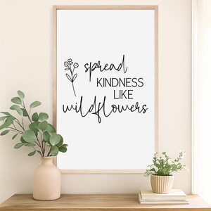 以下が含まれることがあります： 「spread kindness like wildflowers」という言葉が黒い筆記体で書かれた額入りのプリント。プリントは、緑の入った花瓶と小さな鉢植えの植物が置かれた木製のテーブルに飾られています。