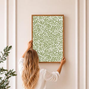 May include: A framed print featuring a repeating pattern of green leaves on a white background. The print is in a wooden frame. The image is being held up against a white wall.