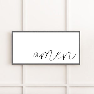 May include: A rectangular wall sign featuring a dark gray frame and a white background. The word amen is written in the center in a black, lowercase, cursive script font. The sign is oriented horizontally and has a clean, simple design.
