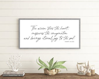 Op de afbeelding: Een houten console met een visgraatpatroon, met decoratieve items. Boven, een ingelijste print met de quote "The ocean stirs the heart..." wordt weergegeven tegen een witte lambrisering. Een kustthema.