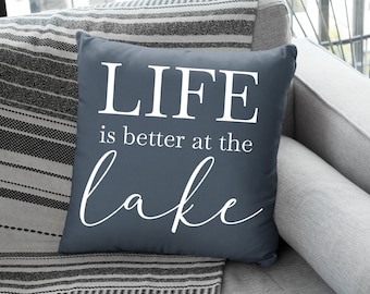 May include: A blue throw pillow with white text that reads "LIFE is better at the lake". The pillow is on a gray couch with a black and white striped throw blanket.