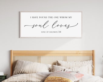 Puede incluir: Una impresión enmarcada blanca con el texto "He encontrado al que ama mi alma. Cantar de los Cantares 3:4" en fuente cursiva negra. La impresión está colgada en una pared blanca sobre una cabecera de cama de madera con un edredón y almohadas beige a rayas.