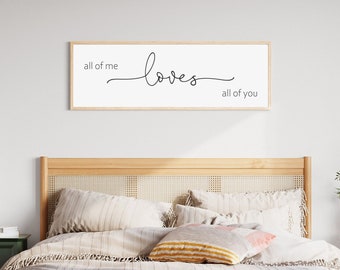 Puede incluir: Una impresión enmarcada en blanco con el texto "all of me loves all of you" colgada sobre una cama con una cabecera de madera y ropa de cama a rayas.