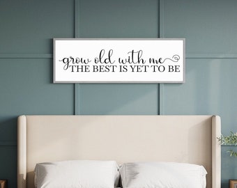 Puede incluir: Un cartel enmarcado en blanco con texto negro que dice "grow old with me ~ the best is yet to be". El cartel está colgado sobre una cama blanca con ropa de cama blanca.