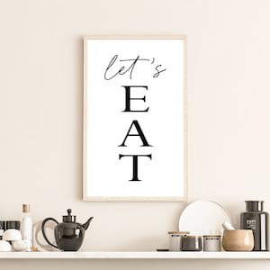 May include: A framed black and white print with the text "let's EAT" in a vertical format. The print is hanging on a white wall above a shelf with various kitchen items.