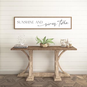 May include: A rectangular wall sign with a light brown wooden frame and a white background featuring the black text SUNSHINE AND river Time. The sign is mounted horizontally on a white wall above a wooden console table with a herringbone top.