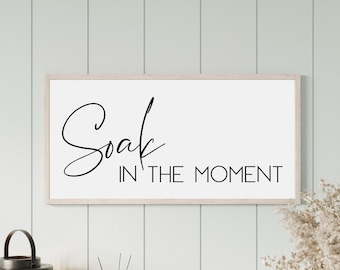 May include: A white framed sign with black calligraphy that reads "Soak in the Moment". The sign is hanging on a light green wall with white vertical wood paneling.