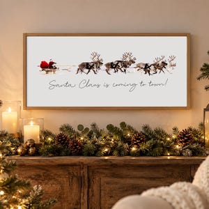 Puede incluir: Un cuadro rectangular con marco de madera clara que presenta un fondo blanco con un gr&aacute;fico de Santa Claus en un trineo tirado por cuatro renos. El texto Santa Claus is coming to town est&aacute; impreso en letra cursiva de color negro.
