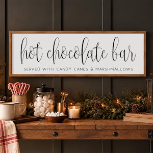 May include: A rectangular wooden sign with a white background and a brown frame featuring the black text "hot chocolate bar" in a script font and "SERVED WITH CANDY CANES & MARSHMALLOWS" in a simple sans-serif font.