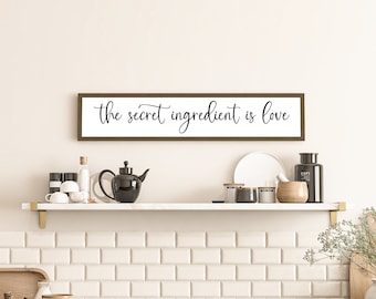 May include: A rectangular wall sign with a dark brown wooden frame and a white background featuring the black cursive text the secret ingredient is love. The sign is mounted horizontally on a plain wall above a white shelf with kitchen items.