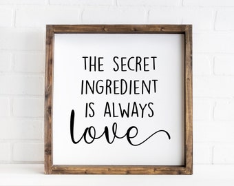 May include: A square wooden sign with a dark brown frame and a white background. The text in black lettering reads: THE SECRET INGREDIENT IS ALWAYS love. The word love is written in a cursive script font, while the other words are in sans-serif.