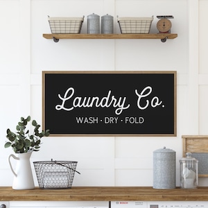 Puede incluir: Un letrero de pared rectangular negro con un marco de madera clara que presenta el texto blanco Laundry Co. y las palabras WASH • DRY • FOLD debajo. El letrero está montado en una pared de paneles blancos sobre un mostrador de madera.